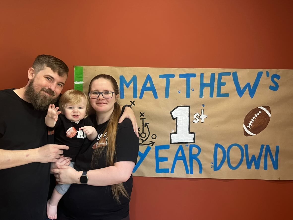 Nick, Libby, and their child standing near a first birthday football sign